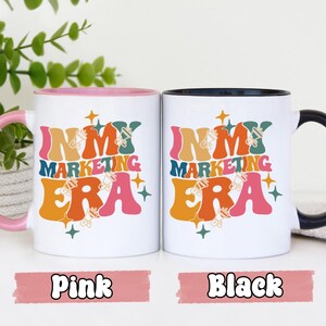 May include: Two white ceramic mugs with black handles. The mugs have a colorful retro design with the text "In My Marketing Era". One mug has a pink handle and the other has a black handle.