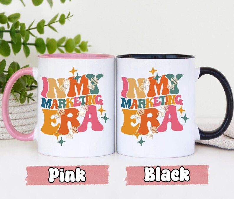 May include: Two white ceramic mugs with black handles. The mugs have a colorful retro design with the text "In My Marketing Era". One mug has a pink handle and the other has a black handle.