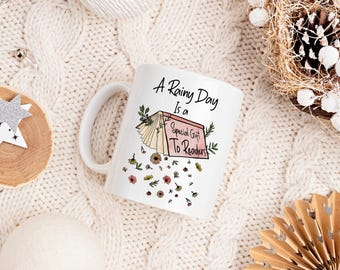 May include: White ceramic mug with a black and white illustration of a book with the text "A Rainy Day Is a Special Gift To Readers". The mug is surrounded by a white knitted fabric.