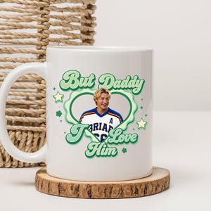 Puede incluir: Taza de cer&aacute;mica blanca con el texto "But Daddy I Love Him" en verde y un dise&ntilde;o en forma de coraz&oacute;n. La taza presenta un retrato de un hombre con una camiseta deportiva. La taza est&aacute; sobre un posavasos de madera.