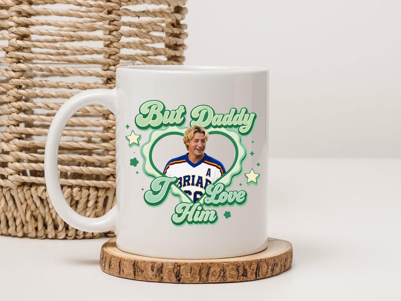 Puede incluir: Taza de cer&aacute;mica blanca con el texto "But Daddy I Love Him" en verde y un dise&ntilde;o en forma de coraz&oacute;n. La taza presenta un retrato de un hombre con una camiseta deportiva. La taza est&aacute; sobre un posavasos de madera.