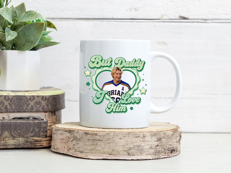 Puede incluir: Taza de cer&aacute;mica blanca con dise&ntilde;o retro. Presenta el texto "But Daddy I Love Him" en verde, una foto de un hombre, estrellas verdes y un marco en forma de coraz&oacute;n.