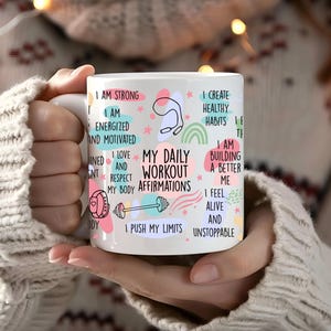 Może przedstawiać: Biały kubek ceramiczny z kolorowymi ilustracjami i tekstem "My Daily Workout Affirmations". Tekst zawiera takie zwroty jak "Jestem silny", "Tworzę zdrowe nawyki", "Przesuwam swoje granice" i "Niezatrzymany".