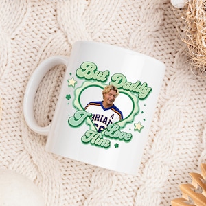 Puede incluir: Taza de cer&aacute;mica blanca con un dise&ntilde;o verde y blanco que dice "But Daddy I Love Him". La taza presenta una foto de un hombre con una camiseta deportiva. La taza tiene un asa blanca y est&aacute; sobre una superficie de punto blanca.