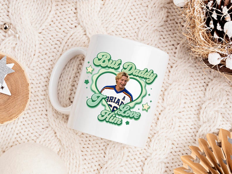 Puede incluir: Taza de cer&aacute;mica blanca con un dise&ntilde;o verde y blanco que dice "But Daddy I Love Him". La taza presenta una foto de un hombre con una camiseta deportiva. La taza tiene un asa blanca y est&aacute; sobre una superficie de punto blanca.