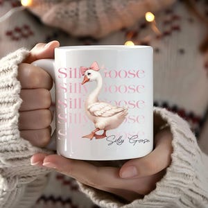 May include: White ceramic mug featuring a whimsical illustration of a goose wearing a pink bow. The mug has the words "Silly Goose" repeated in a pink font. The goose is light pink with orange feet and beak.