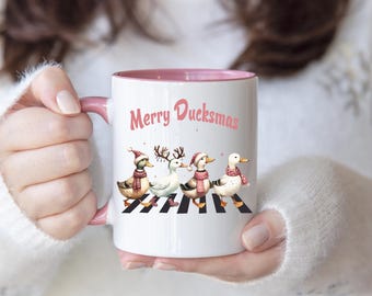 Op de afbeelding: Witte keramische koffiemok met een roze rand. De mok heeft een afbeelding van vier eenden die een kerstmannetje dragen en over een zebrapad lopen. De tekst "Merry Ducksmas" staat op de mok gedrukt.