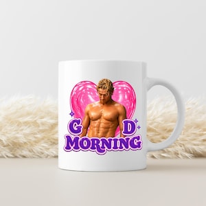 May include: White ceramic mug featuring a muscular man in front of a pink heart. The mug has the words "G MORNING" in purple, stylized text. The design includes a star on each side of the letter "D".