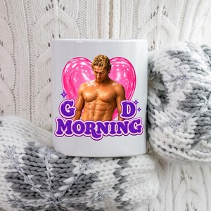 May include: White ceramic mug featuring a muscular man in front of a pink heart. The mug has the words "GOOD MORNING" in purple script. The mug is held by someone wearing a white sweater and gray and white mittens.