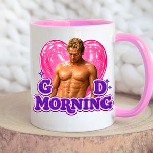May include: White ceramic mug with a pink interior and handle. The mug features a graphic of a muscular person in front of a pink heart, with the words "Good Morning" in purple text.