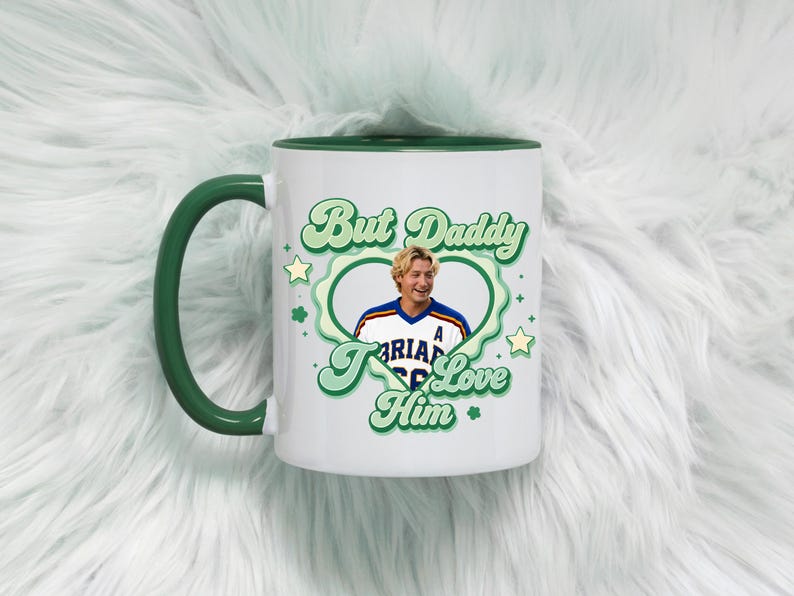 Puede incluir: Taza de cer&aacute;mica blanca con asa y borde verdes. La taza presenta el texto "But Daddy I Love Him" en una fuente verde retro, junto con una foto de un hombre con una camiseta deportiva. La taza est&aacute; sobre una superficie blanca y esponjosa.