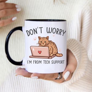 Könnte beinhalten: Weiße Keramiktasse mit schwarzem Rand und Henkel. Die Tasse zeigt eine Comic-Illustration einer Katze, die auf einem Laptop sitzt, mit dem Text "Keine Sorge, ich bin vom technischen Support".