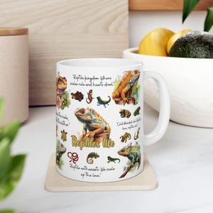 Könnte beinhalten: Eine weiße Keramiktasse mit einer bunten Illustration verschiedener Reptilien, darunter Eidechsen, Schlangen und Schildkröten. Die Tasse zeigt den Text "Reptile kingdom. Where scales rule and hearts drool." und "Reptiles life".
