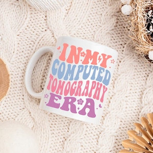 May include: White ceramic mug with a pink, blue, and purple design that says "In My Computed Tomography Era".