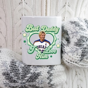 Puede incluir: Taza de cer&aacute;mica blanca con el texto "But Daddy I Love Him" en verde. La taza presenta un retrato de un hombre dentro de un dise&ntilde;o en forma de coraz&oacute;n verde. La taza es sostenida por alguien que lleva un su&eacute;ter blanco y mitones grises.