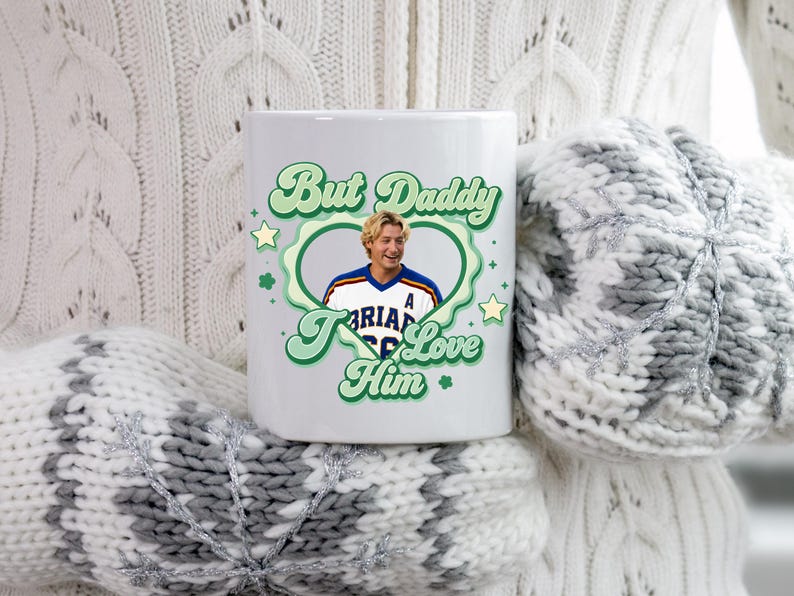 Puede incluir: Taza de cer&aacute;mica blanca con el texto "But Daddy I Love Him" en verde. La taza presenta un retrato de un hombre dentro de un dise&ntilde;o en forma de coraz&oacute;n verde. La taza es sostenida por alguien que lleva un su&eacute;ter blanco y mitones grises.