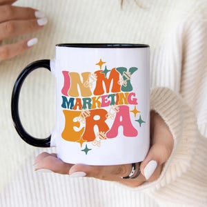 May include: White ceramic mug with a black rim and handle. The mug features a colorful retro-style graphic that says "In My Marketing Era" with stars and other design elements.