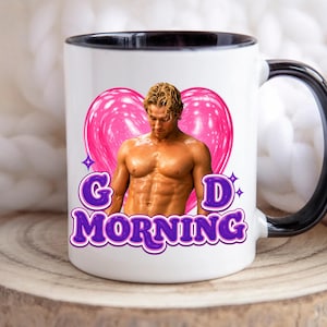 May include: White ceramic mug with a black handle and rim. The mug features a muscular man in front of a pink heart with the words "GOOD MORNING" in purple, outlined in white.
