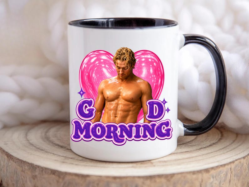 May include: White ceramic mug with a black handle and rim. The mug features a muscular man in front of a pink heart with the words "GOOD MORNING" in purple, outlined in white.