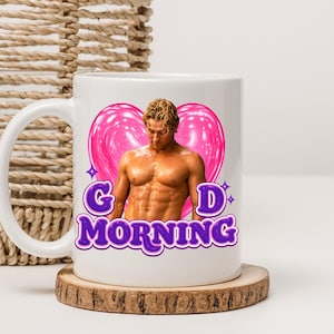 May include: White ceramic mug featuring a muscular man graphic with a pink heart background and the words "G MORNING" in purple. The mug sits on a wooden coaster. A woven basket is in the background.