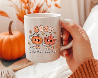 May include: White ceramic mug with a whimsical design featuring a cartoon pumpkin and leaf holding hands, with the words "Cozy Season" in a retro font. The mug is set against a blurred background with a pumpkin and fall foliage.