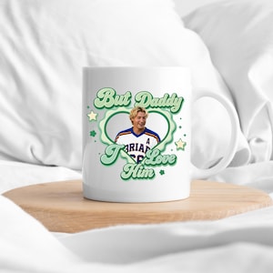 Puede incluir: Taza de cer&aacute;mica blanca con el texto "But Daddy I Love Him" en verde, rodeando una foto de un hombre con una camiseta deportiva. La taza tiene un asa y est&aacute; sobre una superficie de madera.