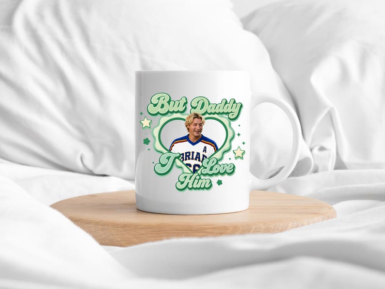 Puede incluir: Taza de cer&aacute;mica blanca con el texto "But Daddy I Love Him" en verde, rodeando una foto de un hombre con una camiseta deportiva. La taza tiene un asa y est&aacute; sobre una superficie de madera.