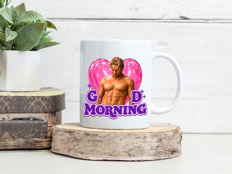 May include: White ceramic mug featuring a muscular man graphic with a pink heart and the words "Good Morning" in purple. The mug is on a wooden slice, with a plant and decorative boxes in the background.
