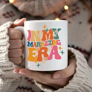 May include: White ceramic mug with a colorful retro design that says "In My Marketing Era". The mug is being held by a person wearing a white sweater.