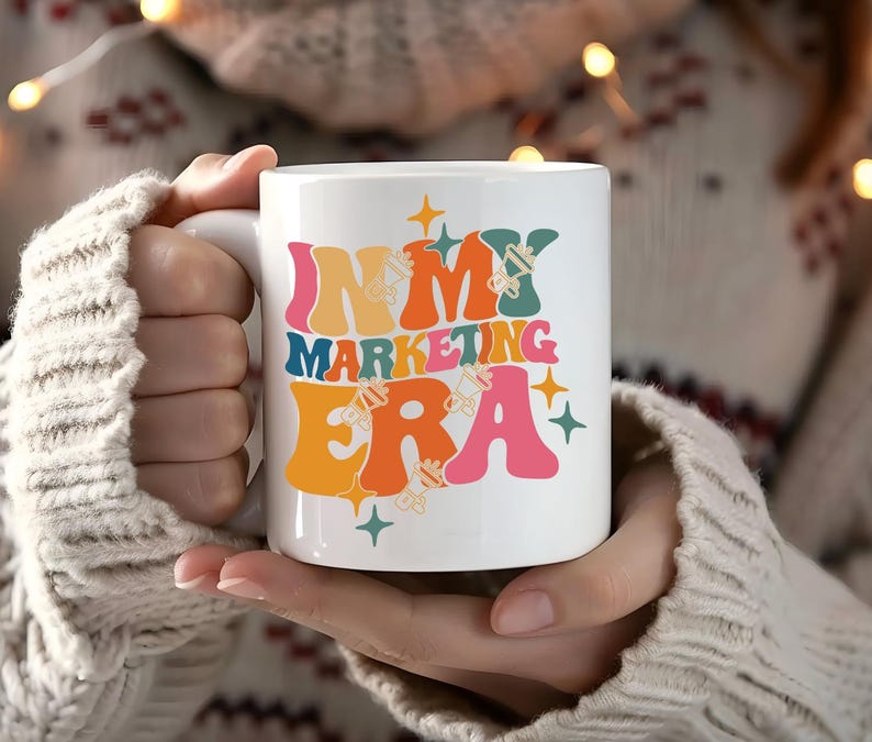 May include: White ceramic mug with a colorful retro design that says "In My Marketing Era". The mug is being held by a person wearing a white sweater.