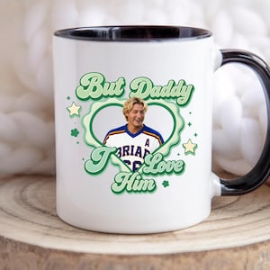 Puede incluir: Taza de cer&aacute;mica blanca con asa y borde negros. La taza presenta un dise&ntilde;o verde y blanco con el texto "But Daddy I Love Him" y una foto de un hombre con una camiseta deportiva. El dise&ntilde;o incluye estrellas y tr&eacute;boles.