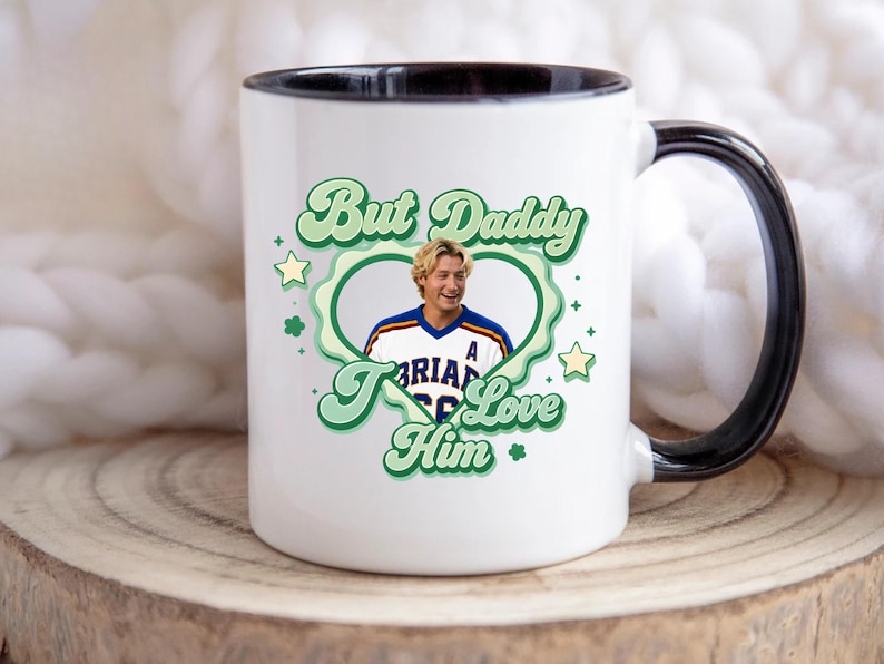 Puede incluir: Taza de cer&aacute;mica blanca con asa y borde negros. La taza presenta un dise&ntilde;o verde y blanco con el texto "But Daddy I Love Him" y una foto de un hombre con una camiseta deportiva. El dise&ntilde;o incluye estrellas y tr&eacute;boles.