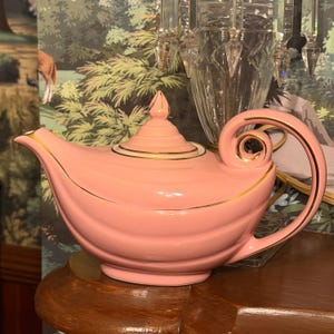 May include: A pastel pink ceramic teapot featuring a curved spout, a matching lid with a pointed finial, and a large, ornate scroll-shaped handle. The teapot is decorated with thin gold trim along the rim, the lid, the spout, and the handle.