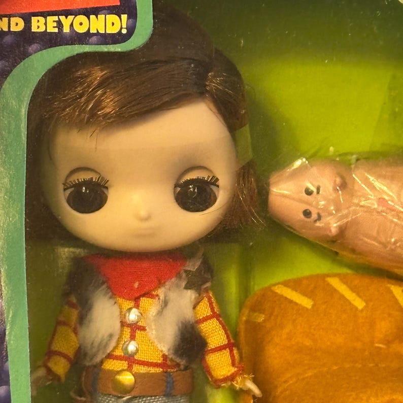 May include: A doll with large eyes, wearing a yellow plaid shirt, a red scarf, and a faux fur vest. The doll is packaged with a pink pig and a yellow hat. The packaging has the text "AND BEYOND!"