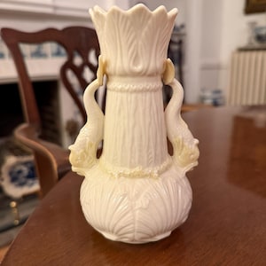 May include: Cream-colored ceramic vase with a flared top and a bulbous base. The vase features two dolphin-shaped handles and a textured, vertical pattern on the body. The top has a scalloped edge, and the vase is set on a wooden table.