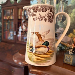 May include: Cream-colored pitcher with a brown floral pattern at the top and a handle. The pitcher features a detailed illustration of ducks in a marshy landscape. A classic piece for serving beverages.