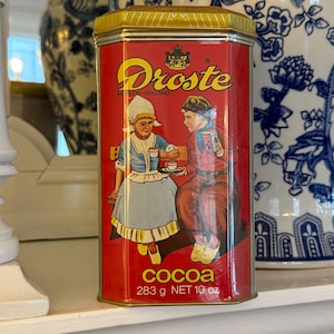 May include: A vintage red cocoa tin with a gold lid and the word "Droste" in a gold script. The tin features an illustration of two people and the text "COCOA 283 g NET 10 oz".