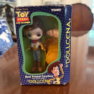 May include: A boxed "Toy Story" Dollcena doll featuring a Woody doll in a cowboy outfit. The box is navy blue with the words "Toy Story" and "Best Friend Cowboy" in English and Japanese. The doll is in a clear plastic window.