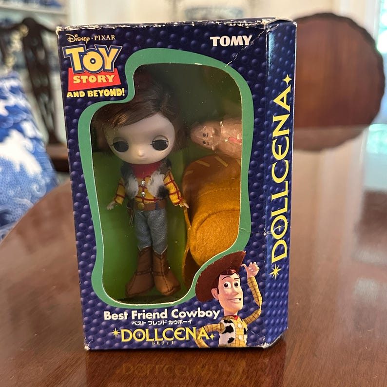 May include: A boxed "Toy Story" Dollcena doll featuring a Woody doll in a cowboy outfit. The box is navy blue with the words "Toy Story" and "Best Friend Cowboy" in English and Japanese. The doll is in a clear plastic window.