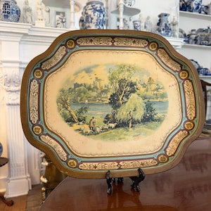 May include: Vintage decorative serving tray with a landscape scene featuring a river, trees, and a castle. The tray has a scalloped edge with a gold and blue border. The tray is supported by a black metal stand.
