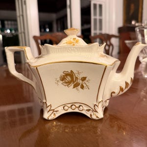 May include: Cream-colored porcelain teapot with gold trim and a floral rose design. The teapot has a square shape with a lid and a curved spout. The handle is also cream-colored with gold accents. A decorative piece for serving tea.