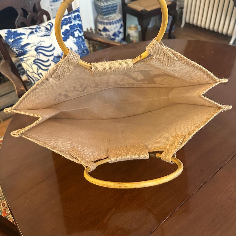 May include: A natural burlap tote bag with bamboo handles. The bag has a rectangular shape with an open top, revealing the interior. The handles are attached with small burlap loops. The bag is designed for carrying items.