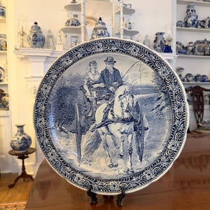 May include: A large, round blue and white decorative plate featuring a detailed illustration of a horse-drawn carriage with two people. The plate has a floral border and is displayed on a wooden table.