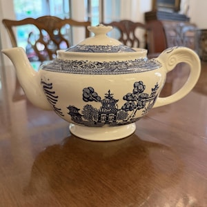 May include: Cream-colored teapot with a blue Willow pattern design. The teapot features a curved spout, a decorative handle, and a matching lid with a small finial. The blue pattern depicts a landscape scene with buildings and trees.