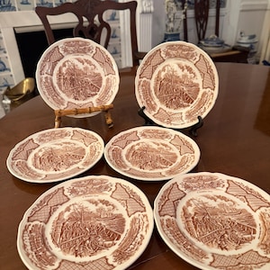 May include: A set of five round ceramic plates featuring a brown and white transferware pattern. Each plate depicts a historical maritime scene with a sailing ship and figures on deck, surrounded by a decorative border with the text A HARBOR VIEW.