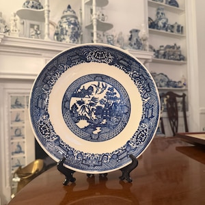 May include: A round ceramic plate featuring a blue and white Willow pattern. The center depicts a landscape with a bridge, trees, and birds, while the wide rim is decorated with a detailed geometric and floral border. The plate rests on a black stand.