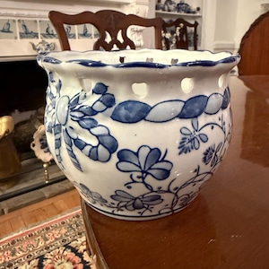 May include: White ceramic planter with blue floral and ribbon designs. The planter has a scalloped rim and small holes around the top. The blue and white color scheme gives it a classic, vintage look. A decorative piece for home decor.