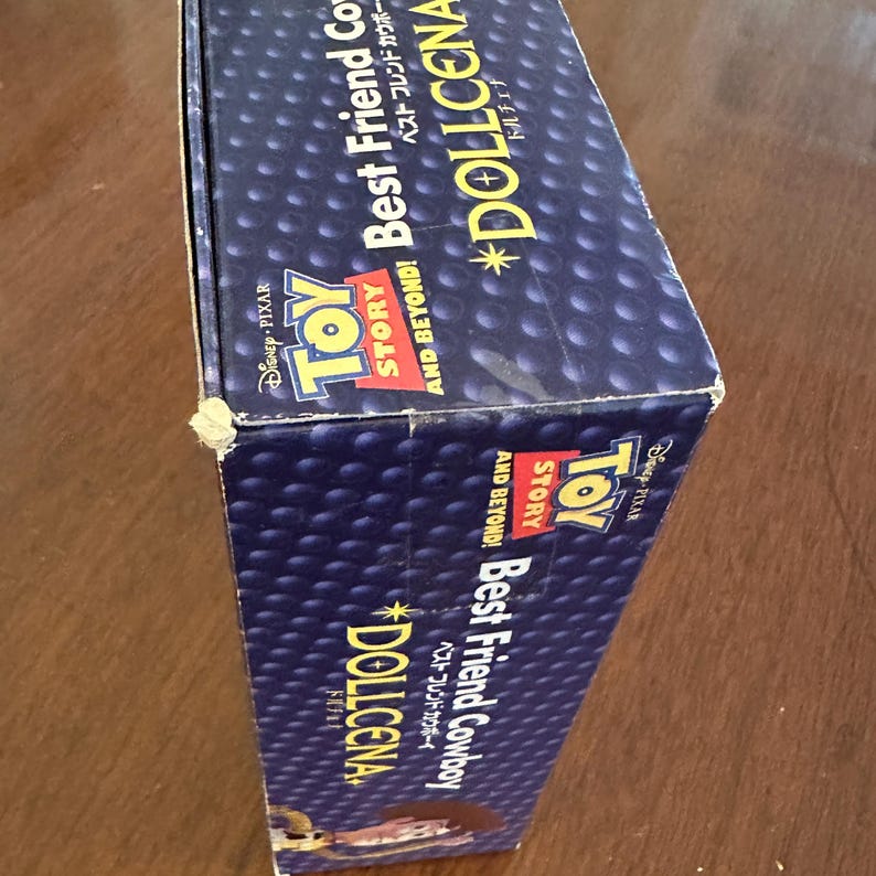 May include: A blue box with the Toy Story logo and the words "Best Friend Cowboy" and "Dollcena" in white and gold. The box has a dotted pattern and the Disney Pixar logo.