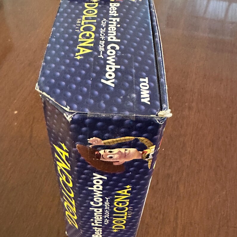 May include: A blue box with the text "Best Friend Cowboy" and "DOLLCENA" in yellow and white. The box features an illustration of a cartoon cowboy. The brand name "TOMY" is also visible.