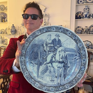 May include: A large round ceramic plate featuring a blue and white transferware design. The central illustration depicts a person driving a horse-drawn carriage with a passenger. The plate has a decorative floral border and scalloped edges.